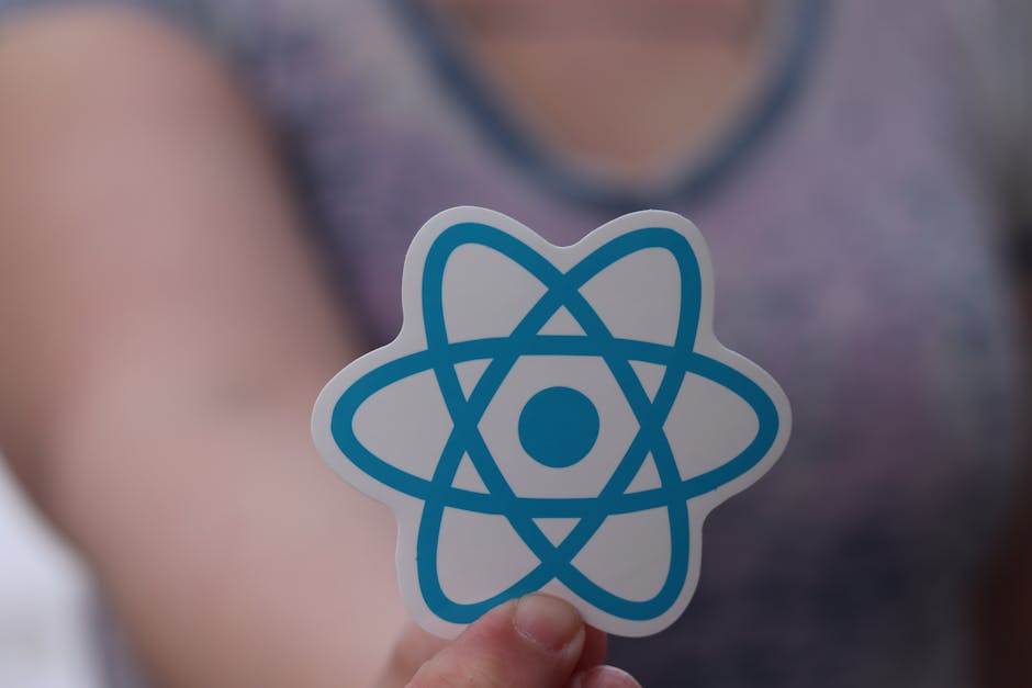 Close-up of a person's hand holding a white sticker with a blue atomic model logo, with the person's blurred face and grey sleeveless top visible in the background. The scene is well-lit, focusing on 